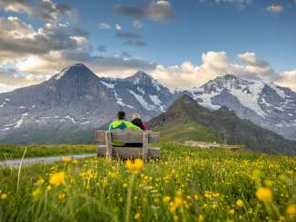 Zwei Wanderer sitzen auf einer Bank. Im Hintergrund Eiger, Mönch und Jungfrau und im Vordergrund eine gelbe Blumenwiese.
